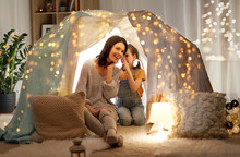 Family, Hygge And People Concept - Happy Mother With Little Daughter Whispering In Kids Tent At Night At Home