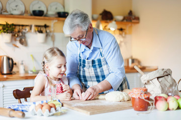 Grandmother is teaching kid to cook pastries and bread in cozy kitchen at home. Senior woman and little girl are happy together. Cute child is helping to prepare dough. Lifestyle authentic moments.