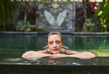  Beautiful real woman relaxing outdoors in lush and exotic stone swimming pool of luxurious resort during tropical vacation in Bali