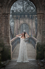 Lovely Young Lady Wearing Elegant White Dress Enjoying The Beams Of Celestial Light And Snowflakes Falling On Her Face. Pretty Brunette Girl In Long Wedding Dress Posing On A Bridge In Winter Scenery