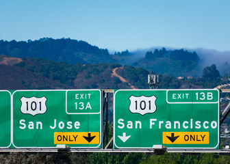 Interstate 101 highway road sign showing drivers the directions to San Jose and San Francisco in Silicon Valley. Green hills with residential area, Dense fog coming from Pacific Ocean in background