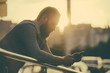 © ruslanshug - beautiful young bearded man in shirt holding cell phone looking at it lit by sunset light beam with flare in urban city background