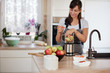 © nenadaksic - Beautiful Caucasian smiling woman in apron putting vegetables and fruits in blender while standing in kitchen.