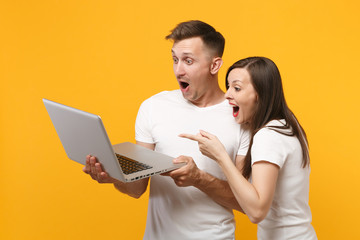 Excited young couple two friends guy girl in white blank t-shirts posing isolated on yellow orange background. People lifestyle concept. Mock up copy space. Holding, working on laptop pc computer.