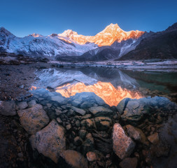 Wall Mural - Snowy mountain with illuminated peaks is reflected in beautiful lake at sunrise. Panoramic landscape with lighted rocks, blue sky, pond, stones in water at dawn. Himalayan mountains in Nepal. Nature