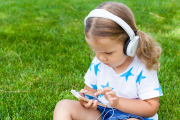 little cute girl sitting in the Park on the grass on a Sunny summer day and listening to music in white headphones. Lifestyle, happy summertime, summer outdoor portrait