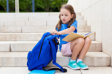 First day of school. child girl schoolgirl elementary school student sitting near the school and puts books in his backpack. Concept back to school. outdoor activities