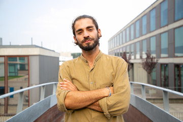 Smiling confident hipster posing for camera in urban settings. Young man in casual with bun and stubble standing with arms crossed and looking at camera. Confident man concept