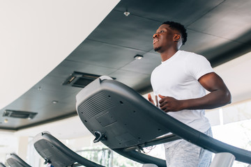 Wall Mural - From below fit African American man running on electric treadmill in gym on daytime. Sportsman working out on training machine