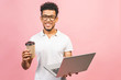 © denis_vermenko - Using laptop. Portrait of handsome casual african american young man drinking coffee while holding laptop computer isolated against pink background.