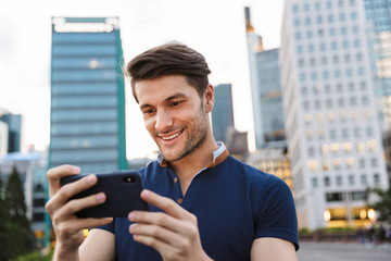 Poster - Pleased cheerful young man walking outdoors by street in city using mobile phone.