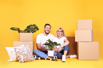 Young married couple moving in new apartment, sitting on the floor between many boxes. New home concept. Bearded man and blonde woman resting over yellow wall. Background, copy space, close up.