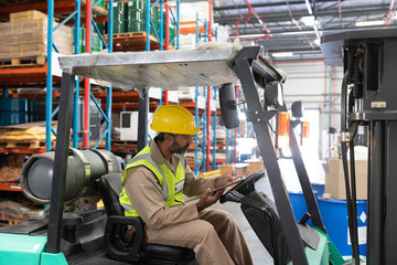 Wall Mural - Male staff writing on clipboard while sitting on forklift in warehouse