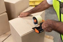 Male Worker Packing Cardboard Box With Tape Gun Dispenser In Warehouse