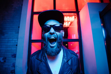 Hipster Handsome Man On The City Streets Being Illuminated By Neon Signs. He Is Wearing Leather Biker Jacket Or Asymmetric Zip Jacket With Black Cap, Jeans And Sunglasses.