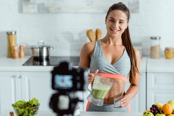 selective focus of happy girl pouring smoothie in glass and looking at digital camera