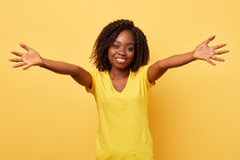 Happy Charming Young Girl With Outstretched Hands Wants To Embrace You Isolated In Yellow Background. Close Up Portrait. Studio Shot.