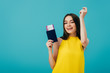 © LIGHTFIELD STUDIOS - cheerful brunette woman in yellow dress dancing with passport with air ticket isolated on turquoise