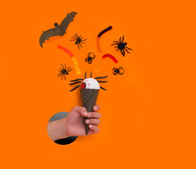 Halloween Holiday Background With Candy Corn And Decorations On An Orange Background. The Children's Hand Holds Ice Cream Cone With Halloween Decor A Hole In Torn Yellow Paper Wall.