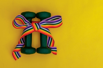 two green dumbbells with rainbow gift bow on yellow table background, pride, sport and healthy concept