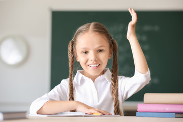 Wall Mural - Cute little girl raising hand during lesson in classroom