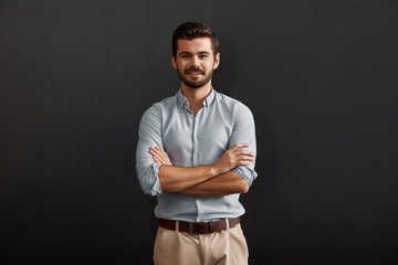 Successful project manager. Cheerful and young bearded man looking at camera with smile and keeping arms crossed while standing against dark background