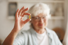 Food, Nutrition, Diet And Health Concept. Close Up Shot Of Elderly Woman's Hand Holding Fish Oil Or Omega-3 Polyunsaturated Fatty Acid Supplement In Shape Of Capsule, Going To Take One During Lunch