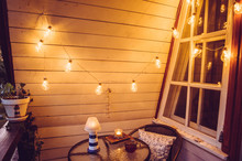 Cute Retro Wooden Nautical Style Balcony View With Small Garden Table And Chair And Decorative String Party Bulbs Lights On In The Evening. 