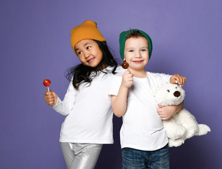 Wall Mural - Kids in colorful hats asian girl and caucasian boy with his polar bear toy posing eating lollipop candy on purple