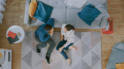 Young Couple is Sitting on a Floor and Talking. They Have an Emotional Dialogue. Cozy Living Room with Modern Interior with Carpet, Sofa, Chair, Table, Book Shelf, Plant and Wooden Floor. Top Down.
