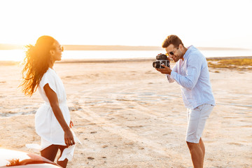 Wall Mural - Image of young man taking photo on retro camera while beautiful woman walking by seaside