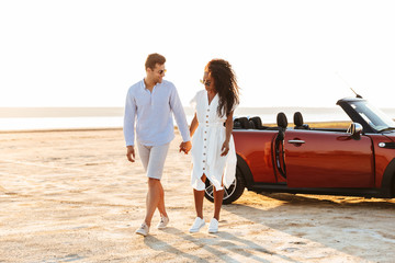 Wall Mural - Photo of young multiethnic couple smiling together while walking by car on beach