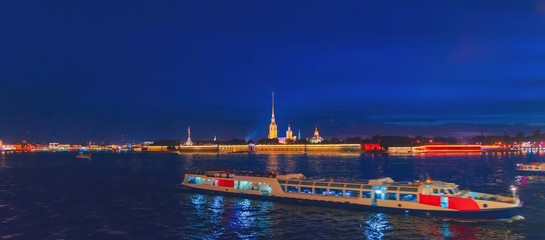 Peter and Paul fortress at night on Neva river with sailing ship, Saint-Petersburg, Russia. Twilight, night lighting illumination, quiet waves river, specular reflection in water, tower spire blue sky