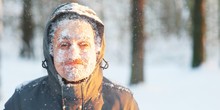 Fun Portrait Of An Young Frozen Man. Jogging In A Blizzard In The Woods. Face Covered With Snow And Frost. Closeup Portrait Of Happy Young Guy Smiles In Cold Weather In The Winter Forest At Sunset.