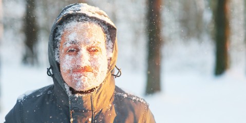 Fun portrait of an young frozen man. Jogging in a blizzard in the woods. Face covered with snow and frost. Closeup portrait of happy young guy smiles in cold weather in the winter forest at sunset.