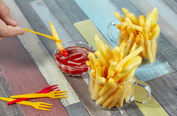 Hand dipping French fries on decorative fork in ketchup. Hand dipping French fries in ketchup. French fries serve in two glass mugs on a wooden surface painted in different colors.