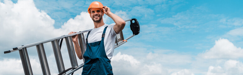 Wall Mural - panoramic shot of cheerful repairman holding ladder and smiling against blue sky with clouds