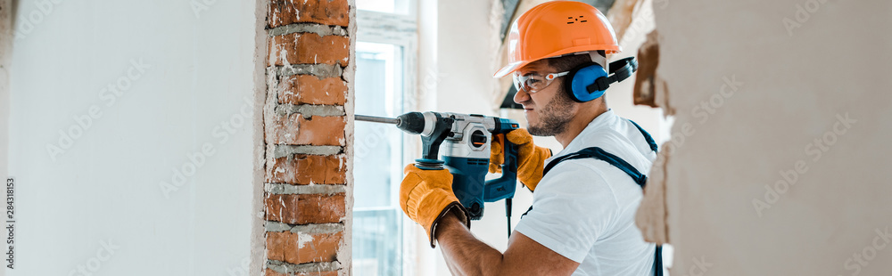 panoramic shot of handyman in uniform and yellow gloves using hammer drill - obrazy, fototapety, plakaty 