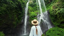 Back of woman in straw hat is enjoying incredible high waterfall Sekumpul (95m) in Bali rainforest. Beautiful wild nature of Indonesia