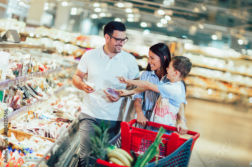 Cuadros en Lienzo Happy family with child and shopping cart buying food at grocery store or superm