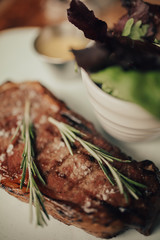 A close up shot of a beef rib eye steak with sea salt and rosemary served on a ceramic plate with salad and sauce. Concept of restaurant, food and culinary.