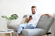 © Evrymmnt - Bearded guy wearing blank white t-shirt & denim pants sitting alone at home on grey textile couch. Young man w/ facial hair in domestic situations. Interior background, copy space, close up, monstera.