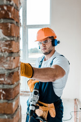 Wall Mural - selective focus of workman in uniform and yellow gloves holding hammer drill and looking at brick wall