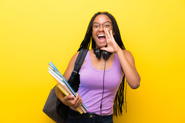 African American teenager student girl with long braided hair over isolated yellow wall shouting with mouth wide open