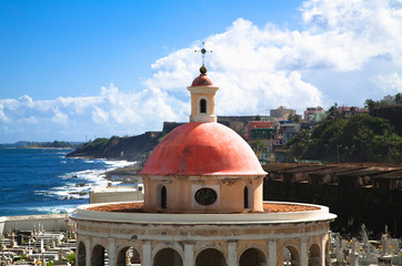 Poster - San Juan, Puerto Rico - An orange dome tops a building in a cemetery. In the background can be seen buildings on a cliff side and the ocean.