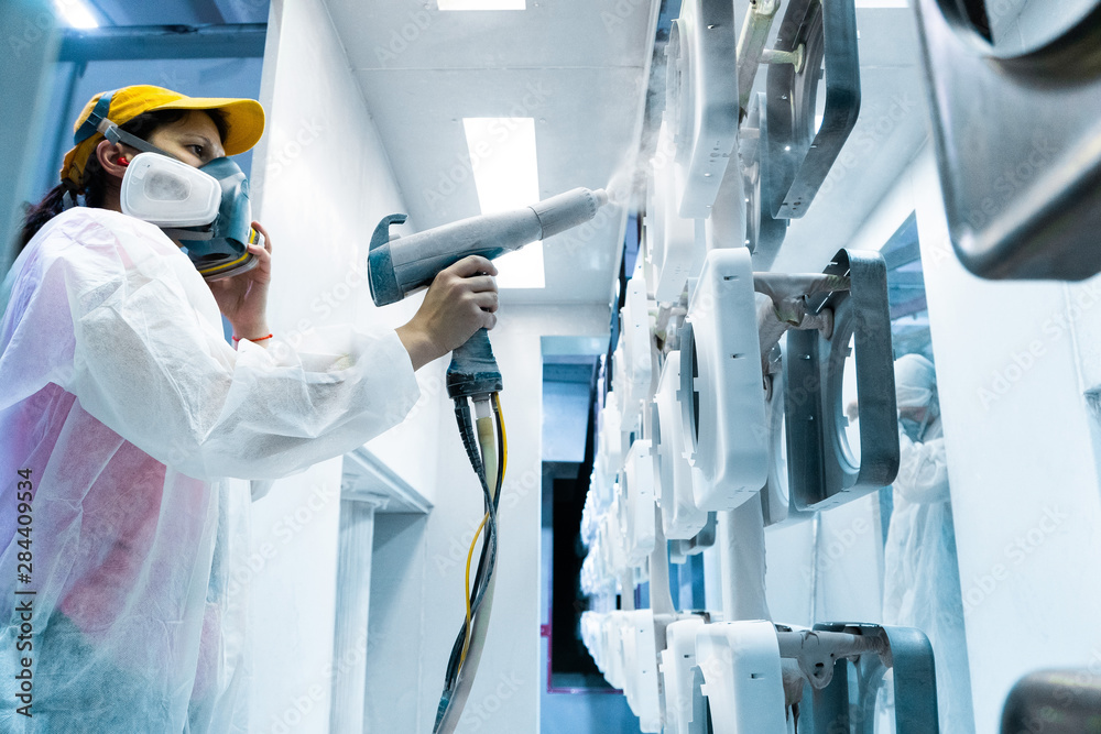 Powder coating of metal parts. A woman in a protective suit sprays white powder paint from a gun on metal products - obrazy, fototapety, plakaty 
