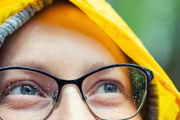 Close-up young tired happy woman portrait wearing glasses with raindrops and bright yellow raincoat hood during rain outdoors