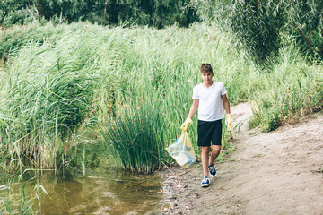 Boy in white t shirt in gloves collects garbage and plastic bottles into blue package on the beach. Young volunteer. Environmental protection, save environment concept