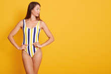 Photo Of Young Woman With Straight Dark Hair Dressed In Swimsuit, Looking Aside Isolated Over Yellow Background In Studio, Keeps Hjands On Hips, Copy Space For Advertismant Or Promotion Text.