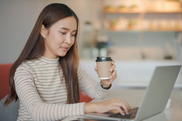 Young beautiful Asian woman working with notebook laptop. She is holding a cup of coffee on hand. Woman smiling and felling happy which looking a notebook at cafe. co working space concept.